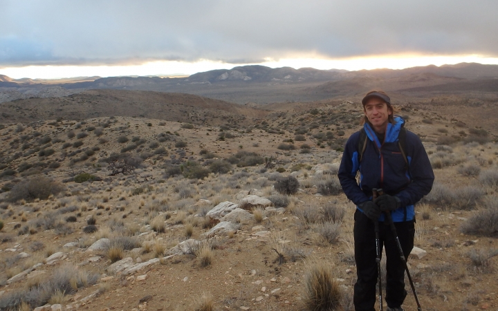 A person wearing a backpack and holding a trekking pole smiles at the camera amidst the desert landscape of Joshua Tree National Park.